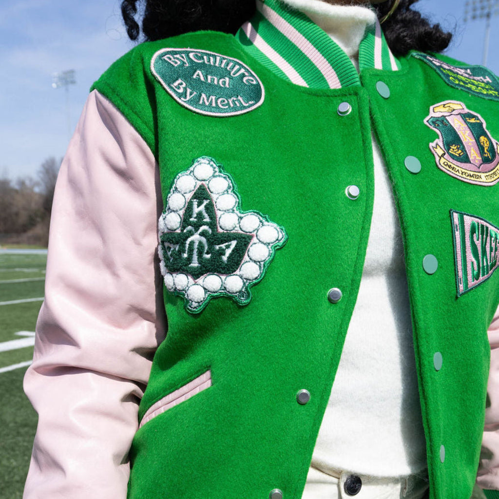 Person wearing a green letterman jacket with various patches on a sports field.