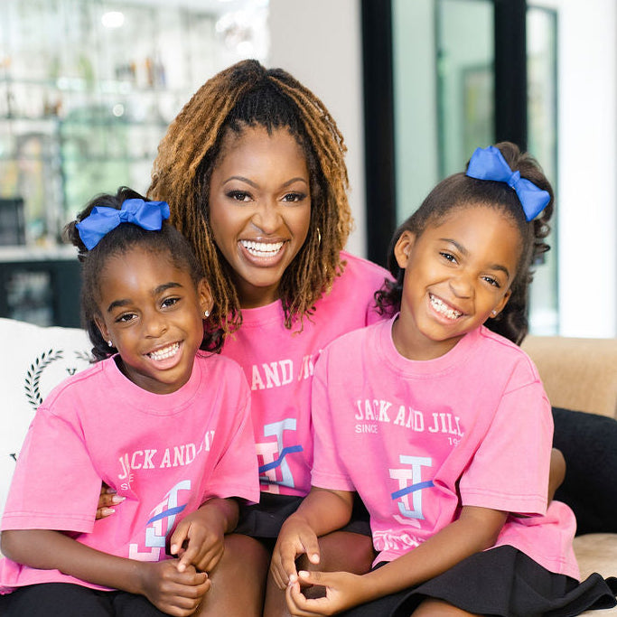 A woman and two young girls sitting together on a beige sofa in a modern living room. The girls are wearing matching pink t-shirts with white text and blue bows in their hair, paired with black skirts and blue sneakers.