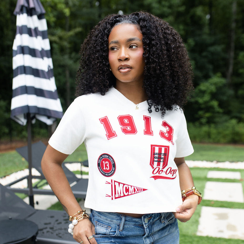 A woman with curly black hair stands outdoors wearing a white cropped V-neck t-shirt with red collegiate-style graphics reading '1913' and distressed blue jeans.