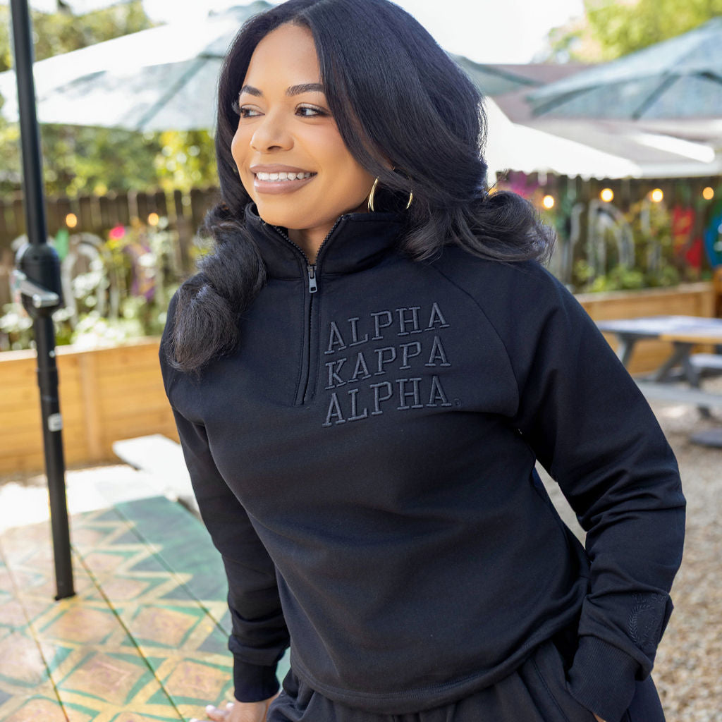 Woman wearing a black quarter zip with 'Alpha Kappa Alpha' text outdoors.