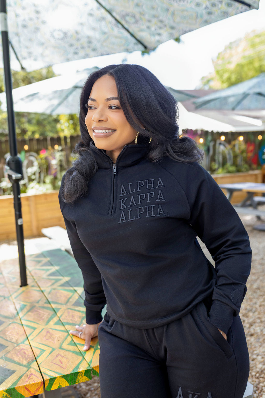 Woman wearing a black quarter zip with 'Alpha Kappa Alpha' text outdoors.