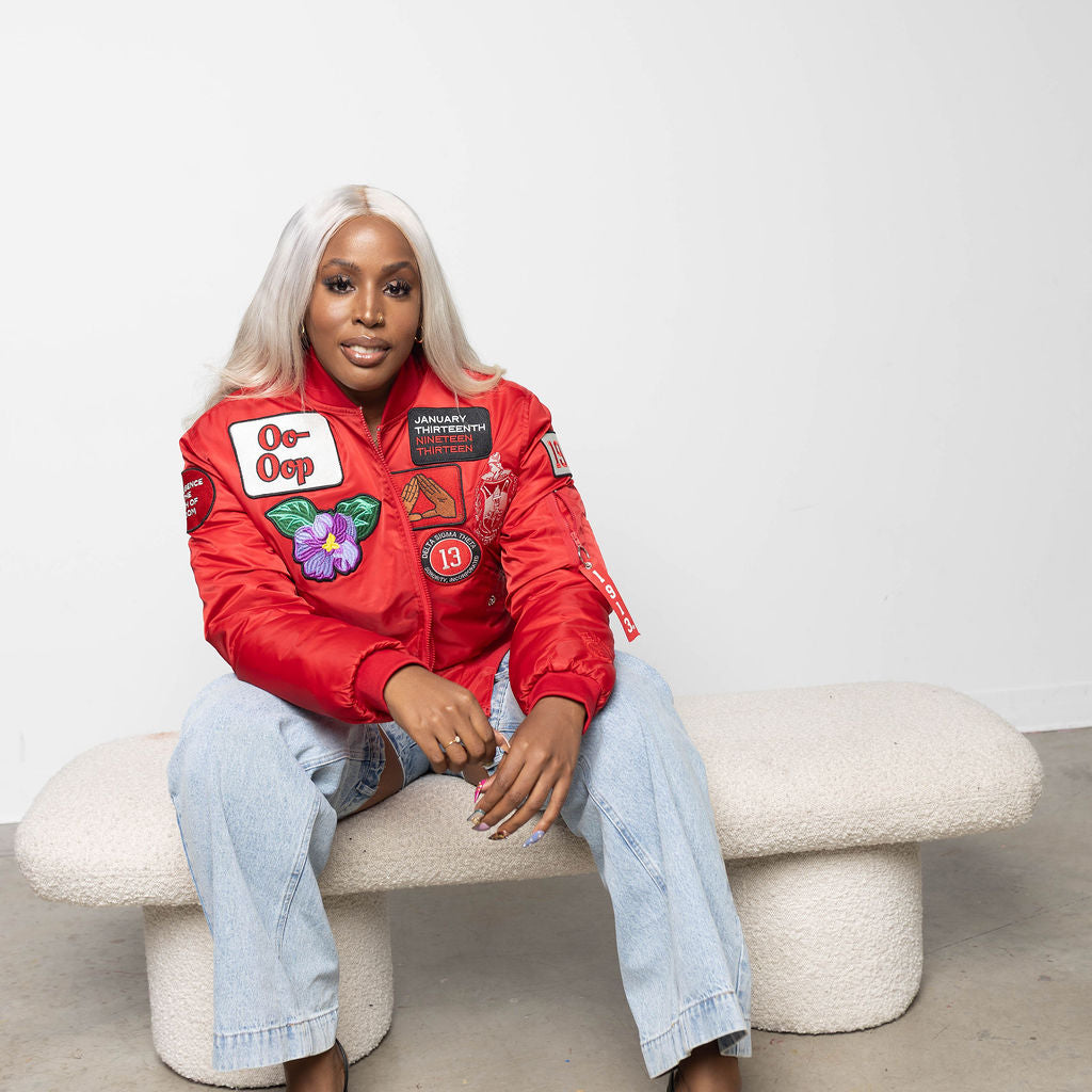 Person wearing a red jacket with delta sigma theta related patches sitting on a white bench against a plain background