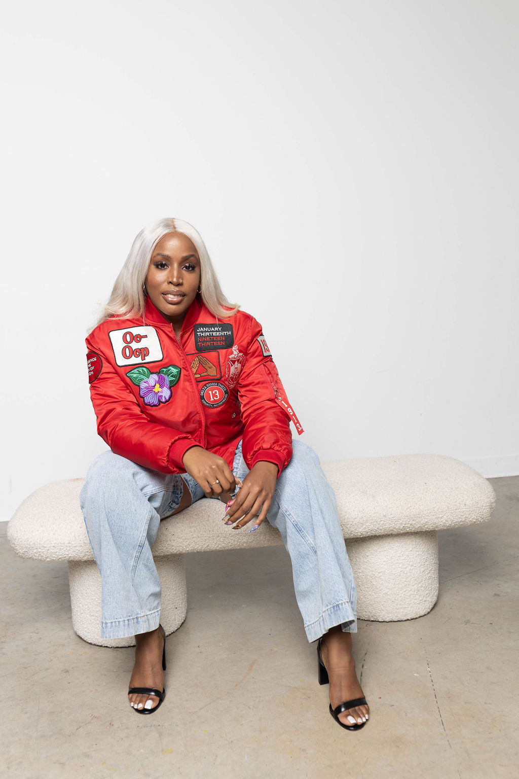 Person wearing a red jacket with delta sigma theta related patches sitting on a white bench against a plain background