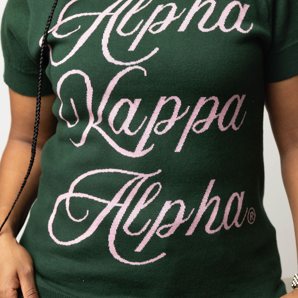 Person wearing a green knit short sleeved sweater with 'Alpha Kappa Alpha' text in pink.