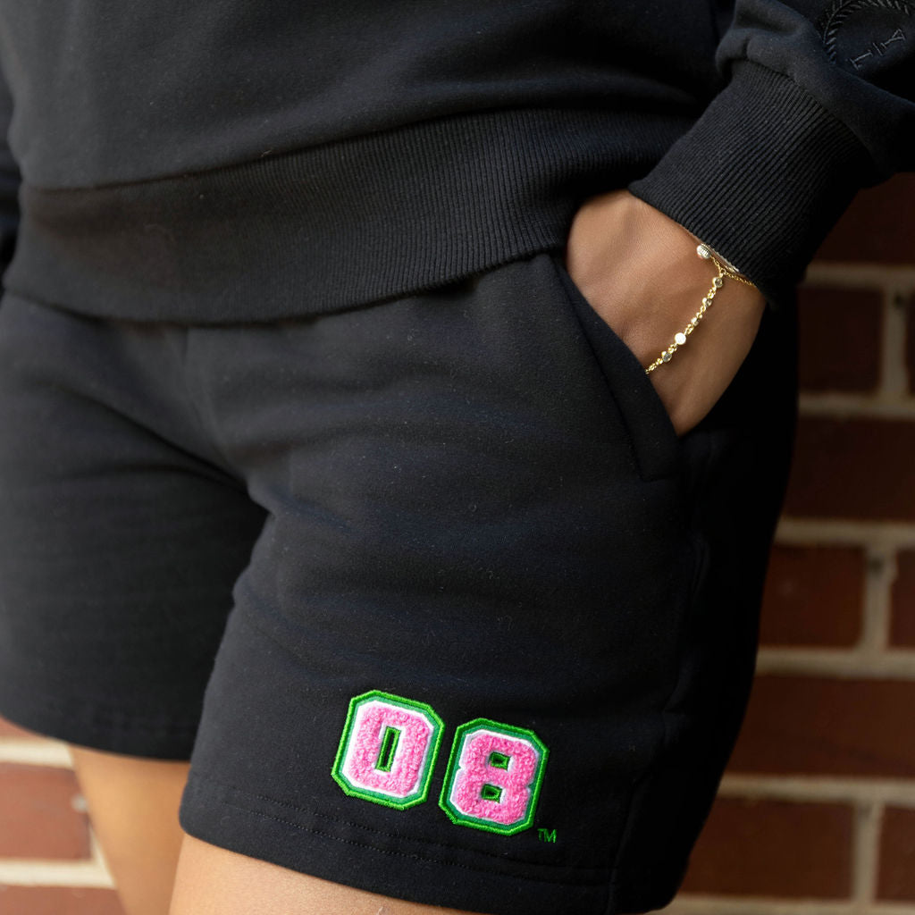 Person wearing black shorts with pink and green 08 design, standing against a brick wall.