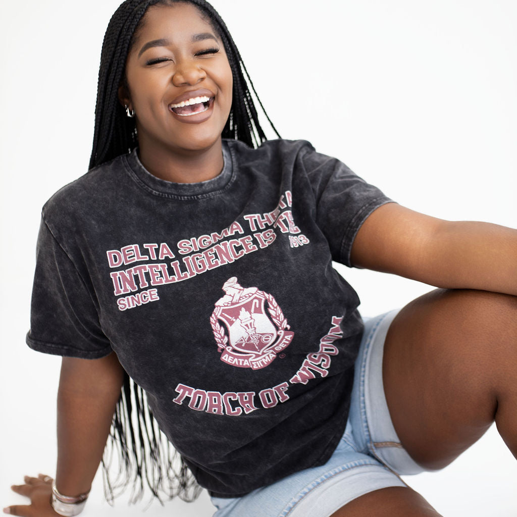 A smiling young woman with long black braids sits on a white studio floor wearing a faded black graphic T-shirt and light denim shorts. The shirt has pink and white text reading 'Delta Sigma Theta' and a crest graphic.
