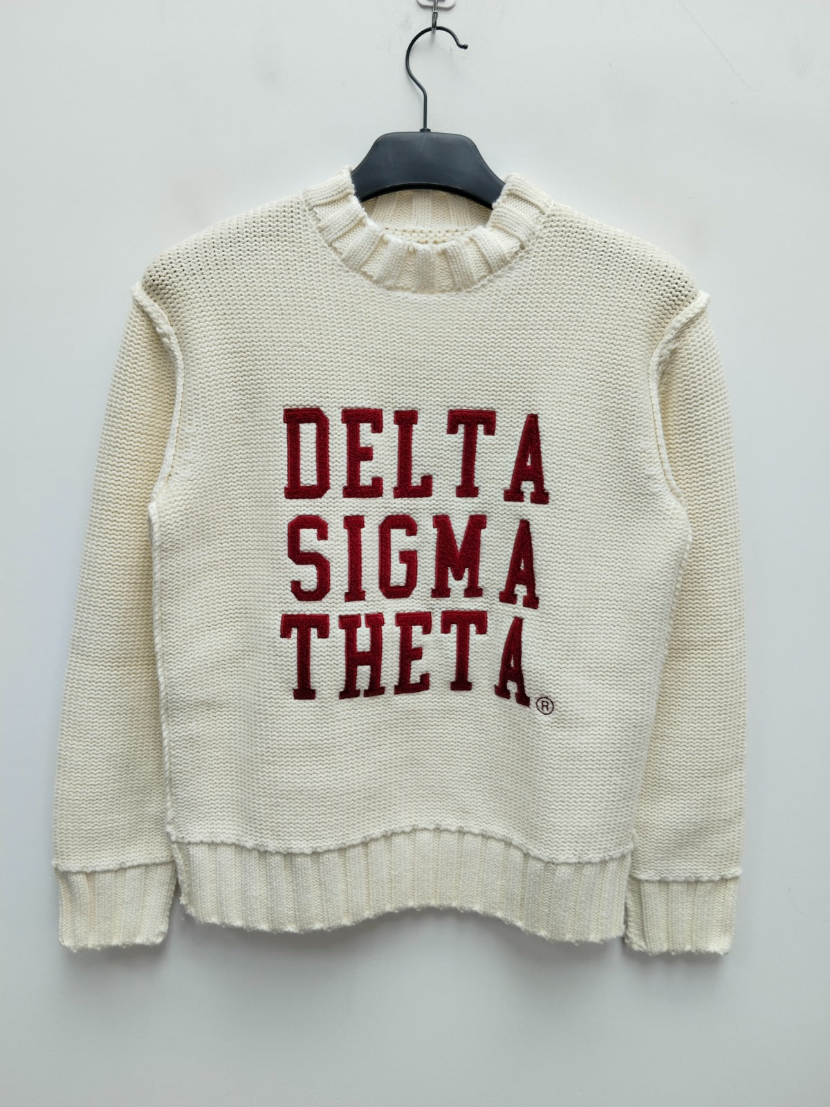 White sweater with 'Delta Sigma Theta' text on a hanger against a light gray background