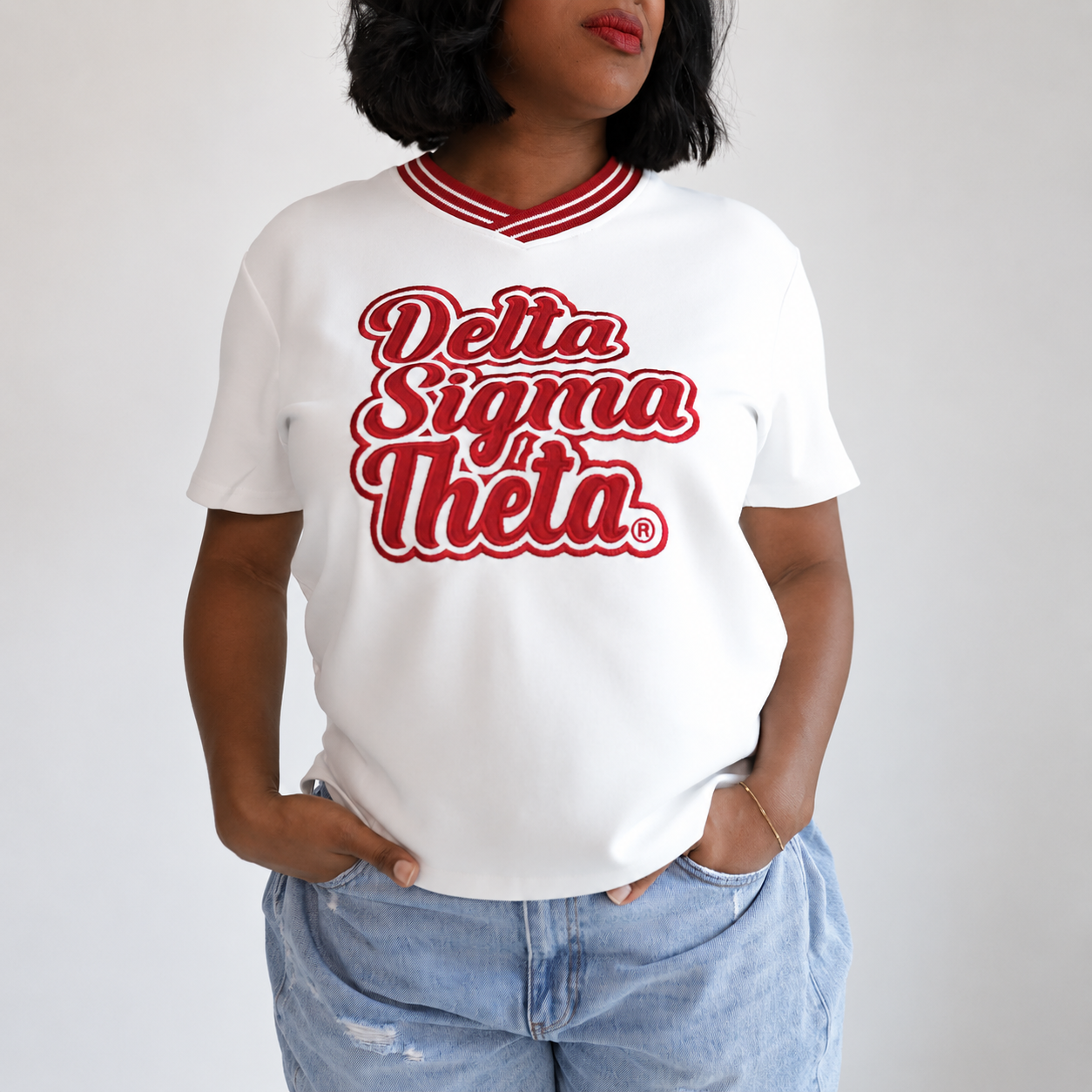A woman wearing a white short-sleeve V-neck T-shirt with 'Delta Sigma Theta' printed in red script across the chest, paired with light blue jeans, against a plain light gray background.