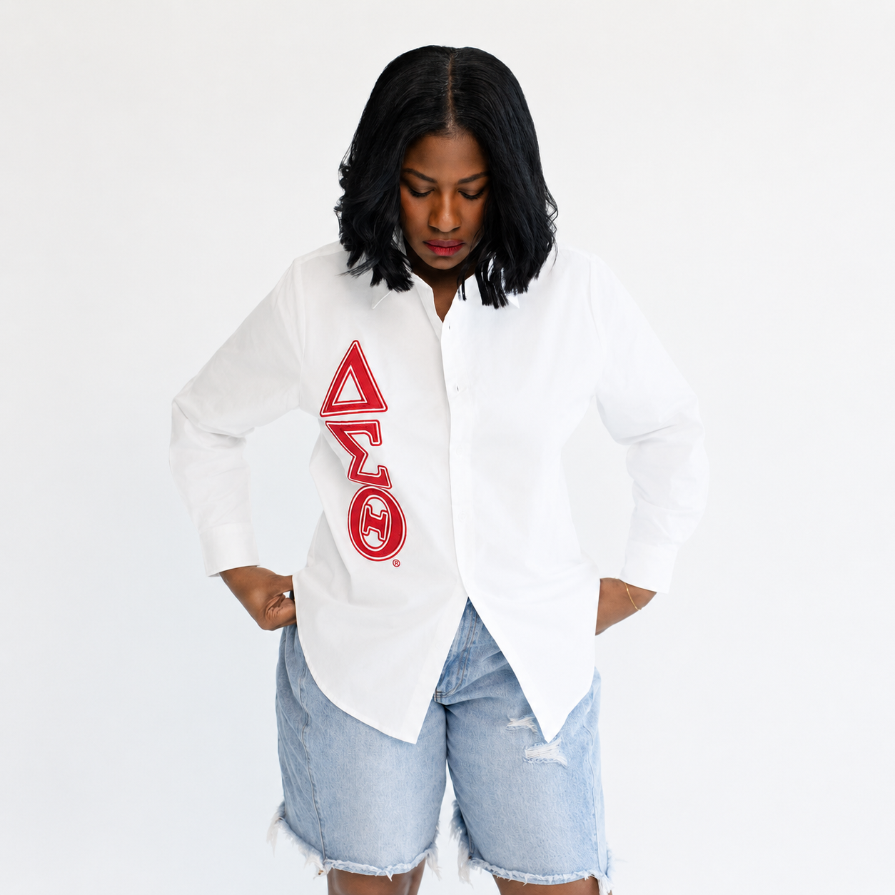A woman wearing a white button-up shirt with a large red Greek letter graphic on the front, paired with light-wash denim shorts, against a plain white background.