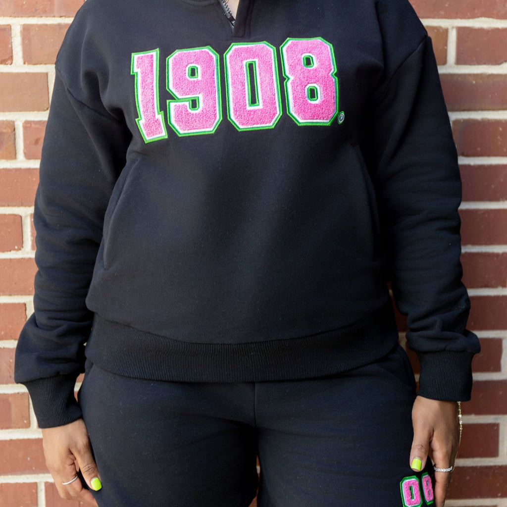 Person wearing a black sweatshirt with '1908' in pink and green letters against a brick wall.