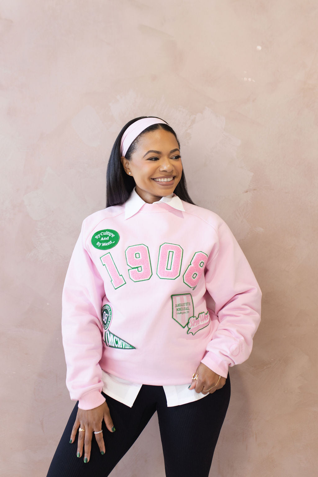Person wearing a pink sweatshirt with text and embroidered patches on a beige background