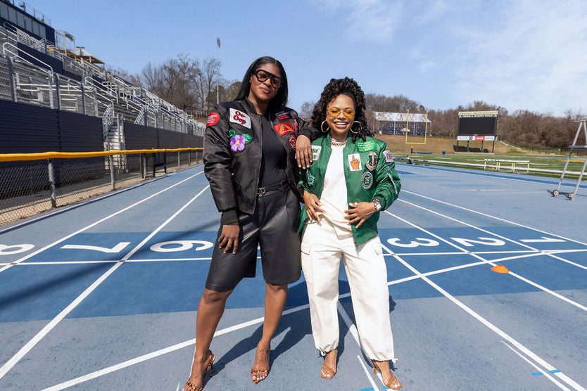 Two women standing on a track field with bleachers and sports equipment in the background.