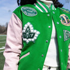 Person wearing a green letterman jacket with various patches on a sports field.