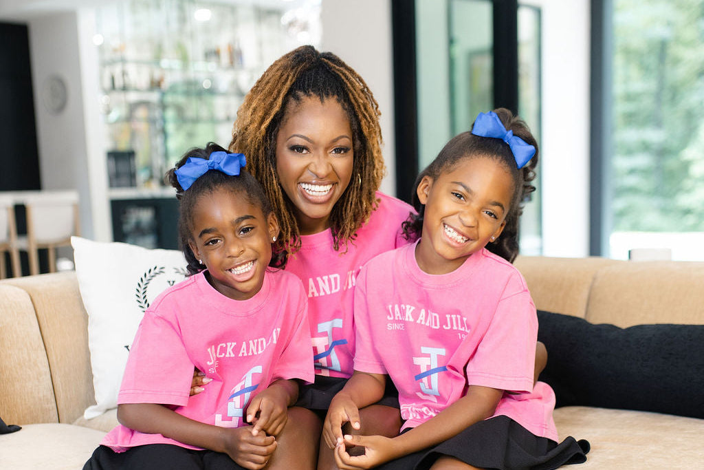 A woman and two young girls sitting together on a beige sofa in a modern living room. The girls are wearing matching pink t-shirts with white text and blue bows in their hair, paired with black skirts and blue sneakers.
