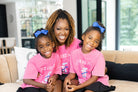 A woman and two young girls sitting together on a beige sofa in a modern living room. The girls are wearing matching pink t-shirts with white text and blue bows in their hair, paired with black skirts and blue sneakers.