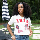 A woman with curly black hair stands outdoors wearing a white cropped V-neck t-shirt with red collegiate-style graphics reading '1913' and distressed blue jeans.