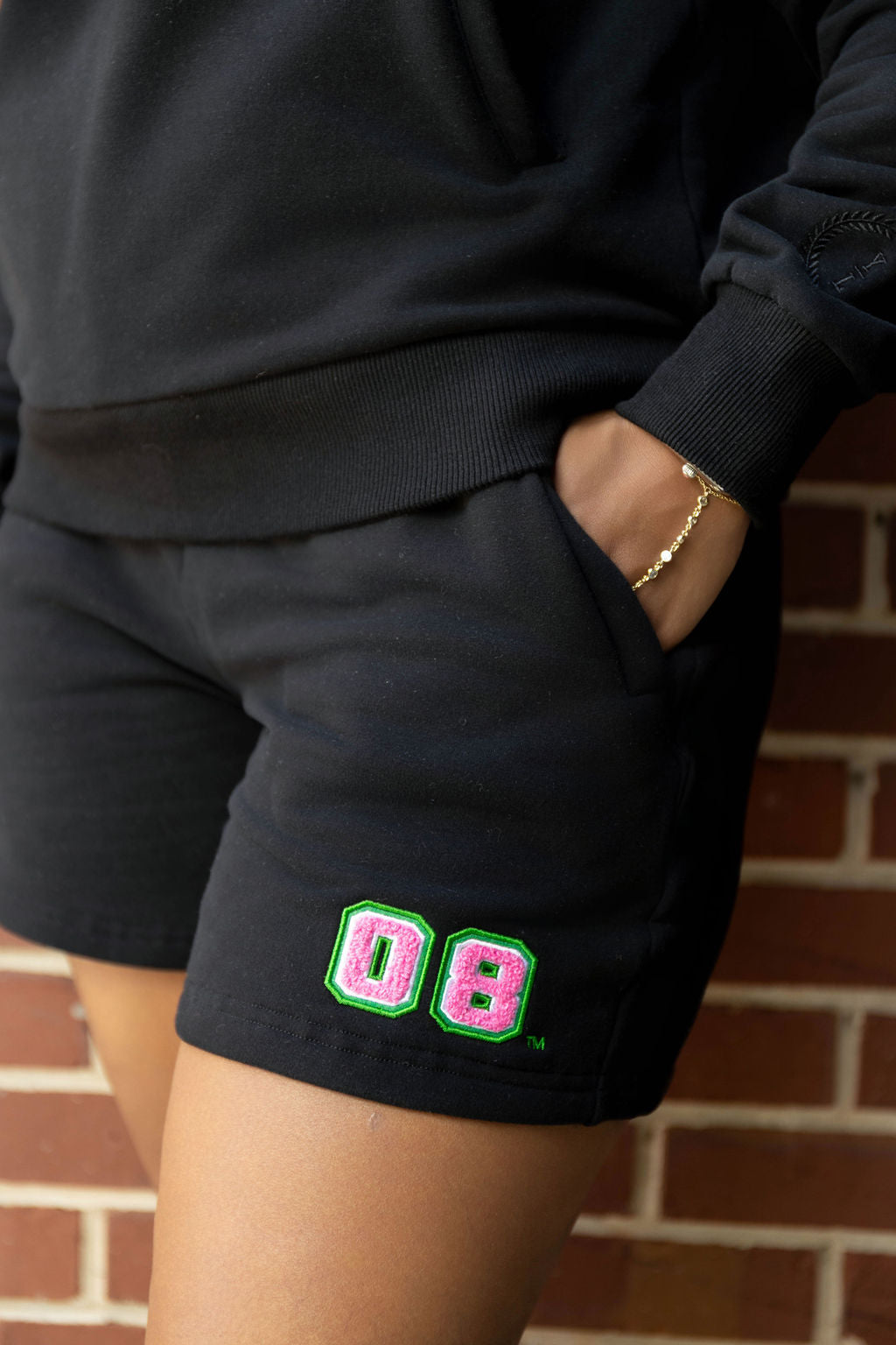 Person wearing black shorts with pink and green 08 design, standing against a brick wall.