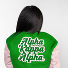 Person wearing a green 'Alpha Kappa Alpha' letterman jacket on a white background