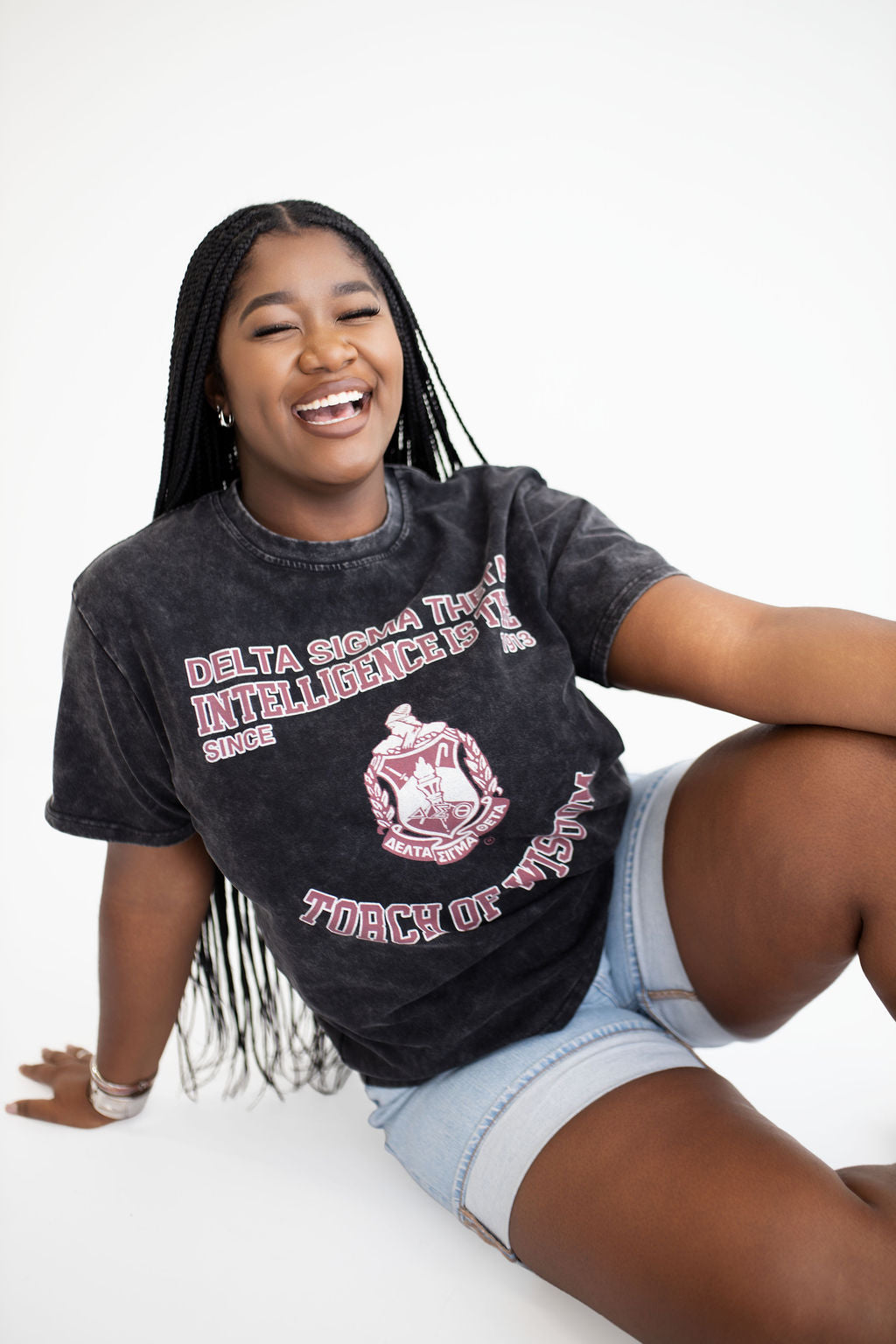 A smiling young woman with long black braids sits on a white studio floor wearing a faded black graphic T-shirt and light denim shorts. The shirt has pink and white text reading 'Delta Sigma Theta' and a crest graphic.