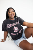 A smiling young woman with long black braids sits on a white studio floor wearing a faded black graphic T-shirt and light denim shorts. The shirt has pink and white text reading 'Delta Sigma Theta' and a crest graphic.