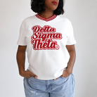A woman wearing a white short-sleeve V-neck T-shirt with 'Delta Sigma Theta' printed in red script across the chest, paired with light blue jeans, against a plain light gray background.