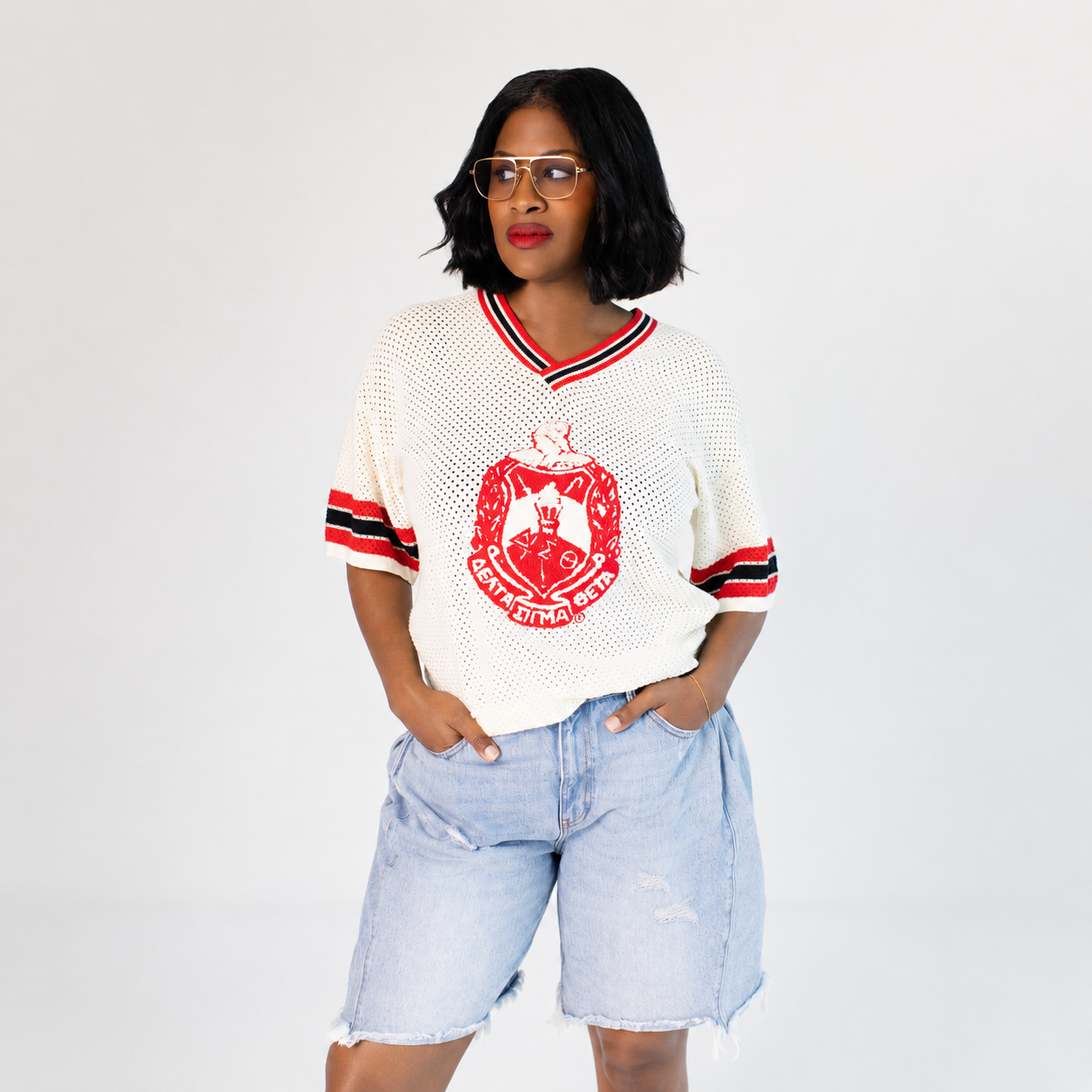A woman modeling a white knit V-neck delta sigma theta jersey-style top with red and black striped trim, a large red crest graphic on the front, and red-and-black striped sleeve cuffs, paired with light blue denim shorts against a light gray studio background.
