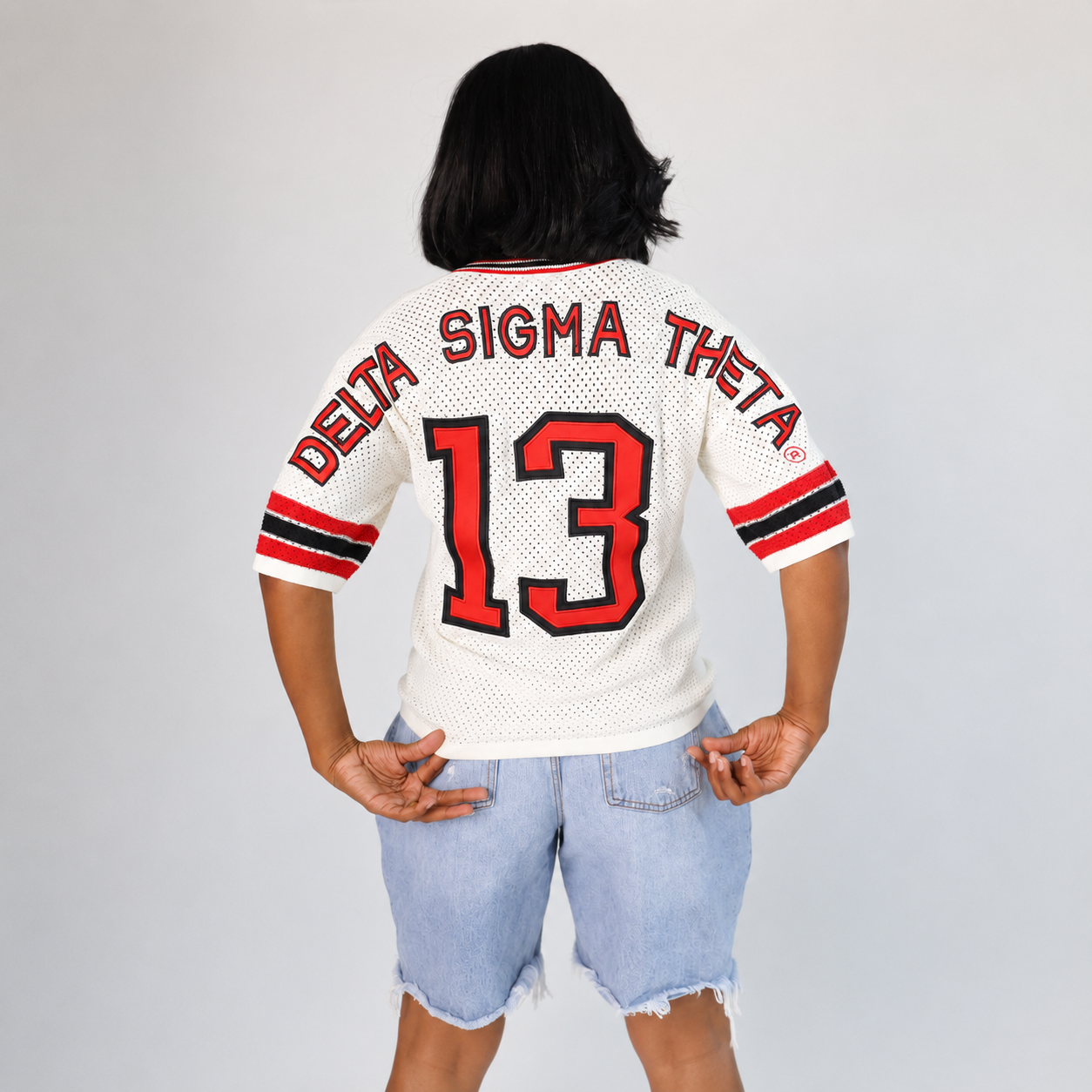 A person shown from the back wearing a white mesh football-style jersey with red and black trim, 'DELTA SIGMA THETA' across the shoulders, and the number 13 on the back. The person is also wearing light blue denim shorts against a plain light gray studio background.