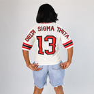 A person shown from the back wearing a white mesh football-style jersey with red and black trim, 'DELTA SIGMA THETA' across the shoulders, and the number 13 on the back. The person is also wearing light blue denim shorts against a plain light gray studio background.