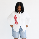 A woman wearing a white button-up shirt with a large red Greek letter graphic on the front, paired with light-wash denim shorts, against a plain white background.