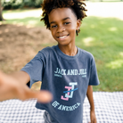 A smiling young boy wearing a navy blue graphic t-shirt with 'JACK AND JILL OF AMERICA' printed on the front, standing outdoors on a checkered blanket.