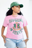 A woman wearing a pink graphic t-shirt with green Greek lettering and a crest, paired with a green baseball cap and denim shorts, against a plain white background.