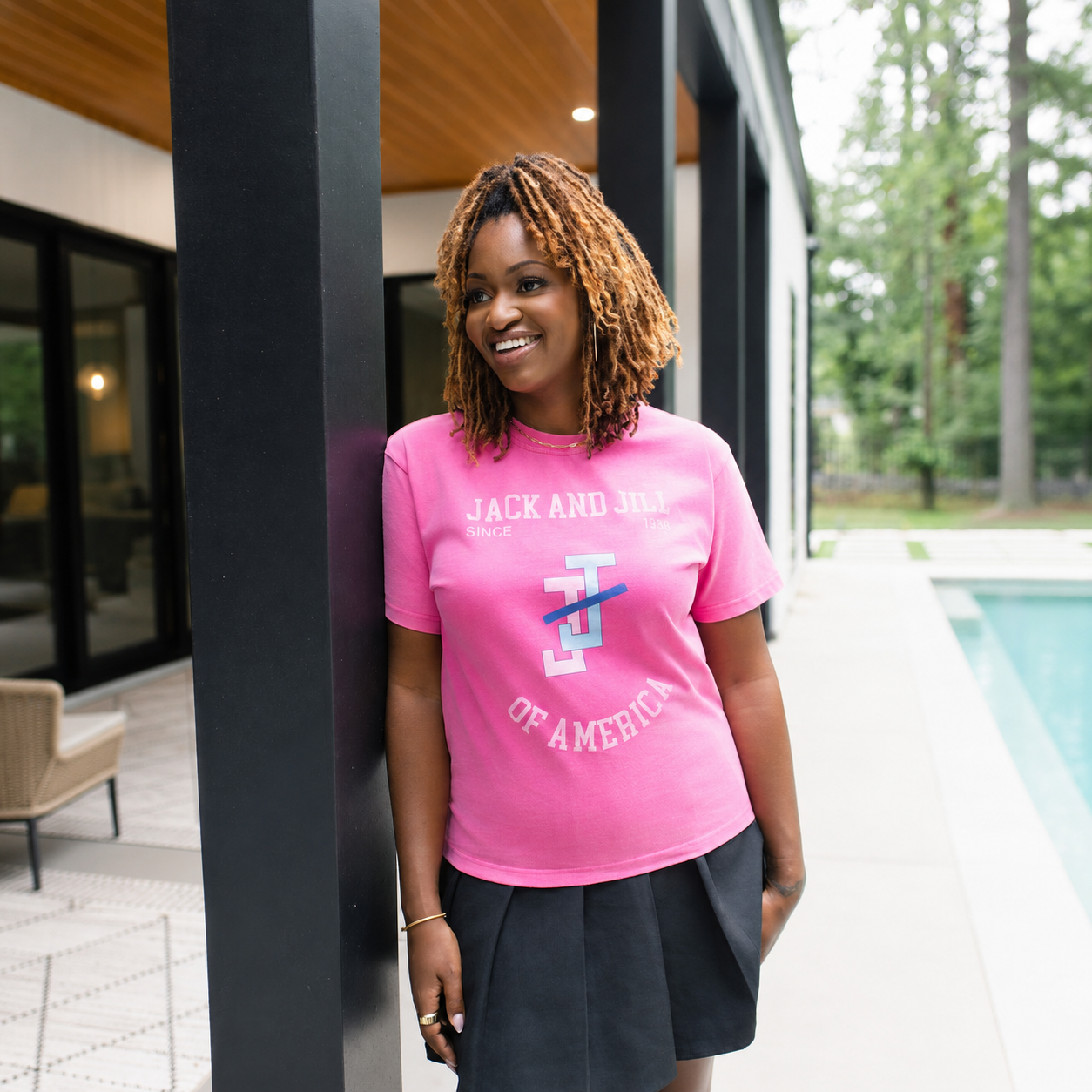 A woman with blonde dreadlocks stands on a patio next to a pool wearing a bright pink graphic t-shirt with 'JACK AND JILL' and 'OF AMERICA' text, paired with a dark skirt.