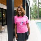 A woman with blonde dreadlocks stands on a patio next to a pool wearing a bright pink graphic t-shirt with 'JACK AND JILL' and 'OF AMERICA' text, paired with a dark skirt.