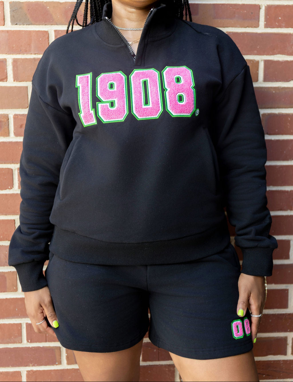 Person wearing a black sweatshirt with '1908' in pink and green letters against a brick wall.