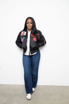 Person wearing a black bomber jacket with patches, white shirt, blue jeans, and white sneakers on a plain background