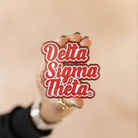 Person holding a 'Delta Sigma Theta' pin against a beige background